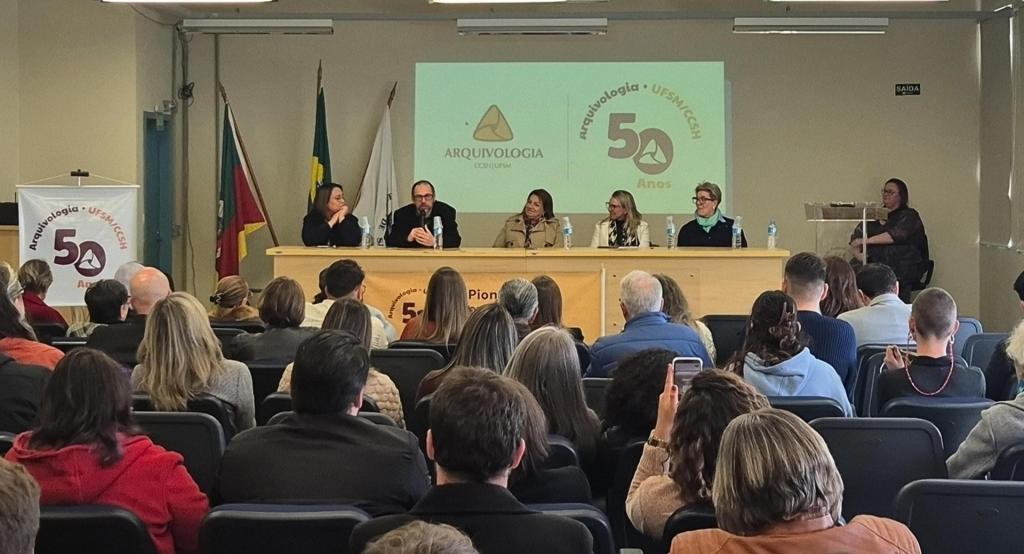 Foto colorida horizontal de cinco pessoas sentadas na mesa de abertura do evento, incluido a reitora e as demais autoridades. À frente, o auditório lotado de ouvintes.