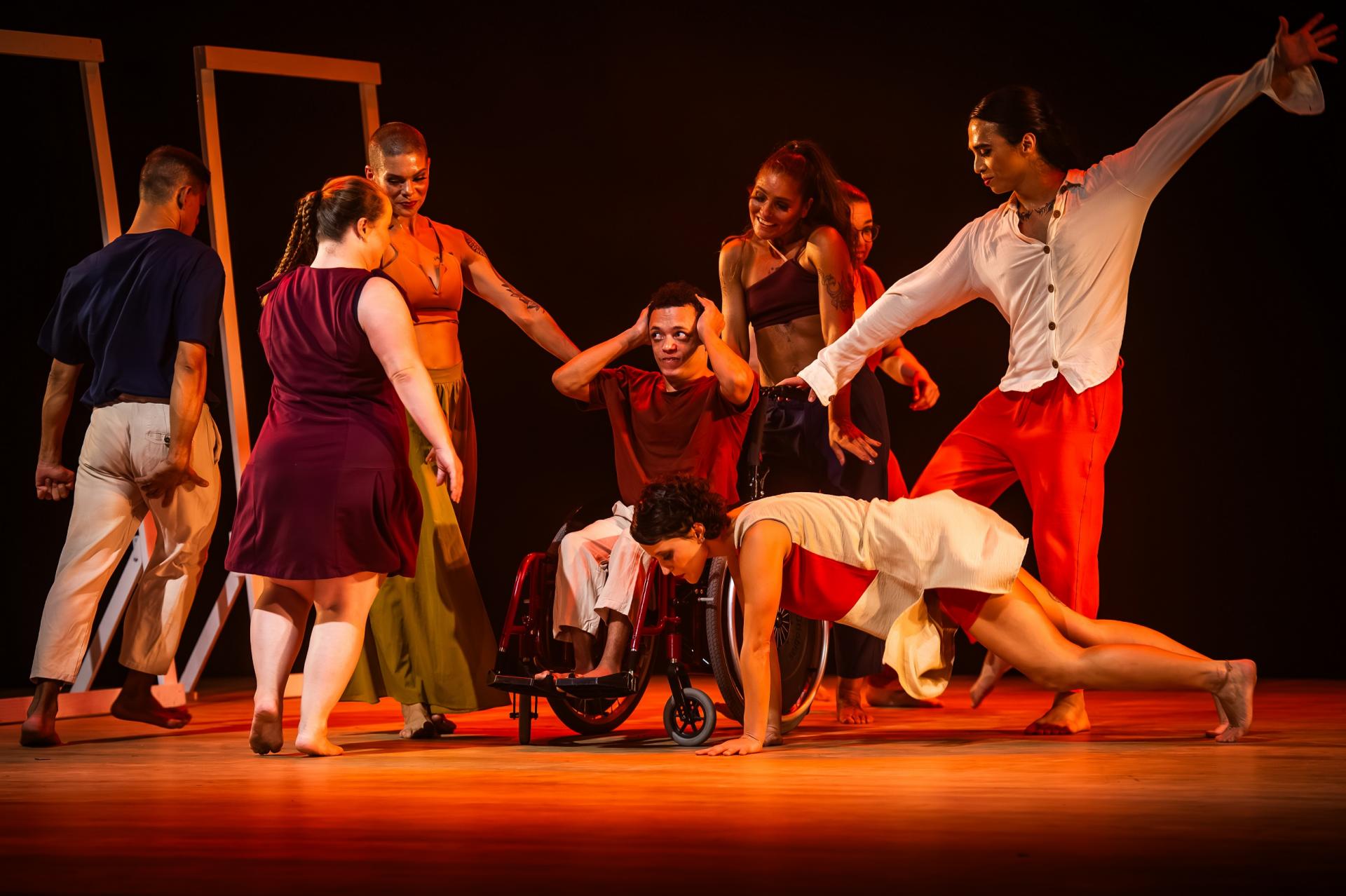 Foto colorida horizontal de oito dançarinos no palco, entre mulheres e homens. Seis estão em pé, alguns de frente e outros de costas, um em uma cadeira de rodas, e a oitava bailarina está em posição horizontal apoiada sobre os branços e as pernas flexionadas.