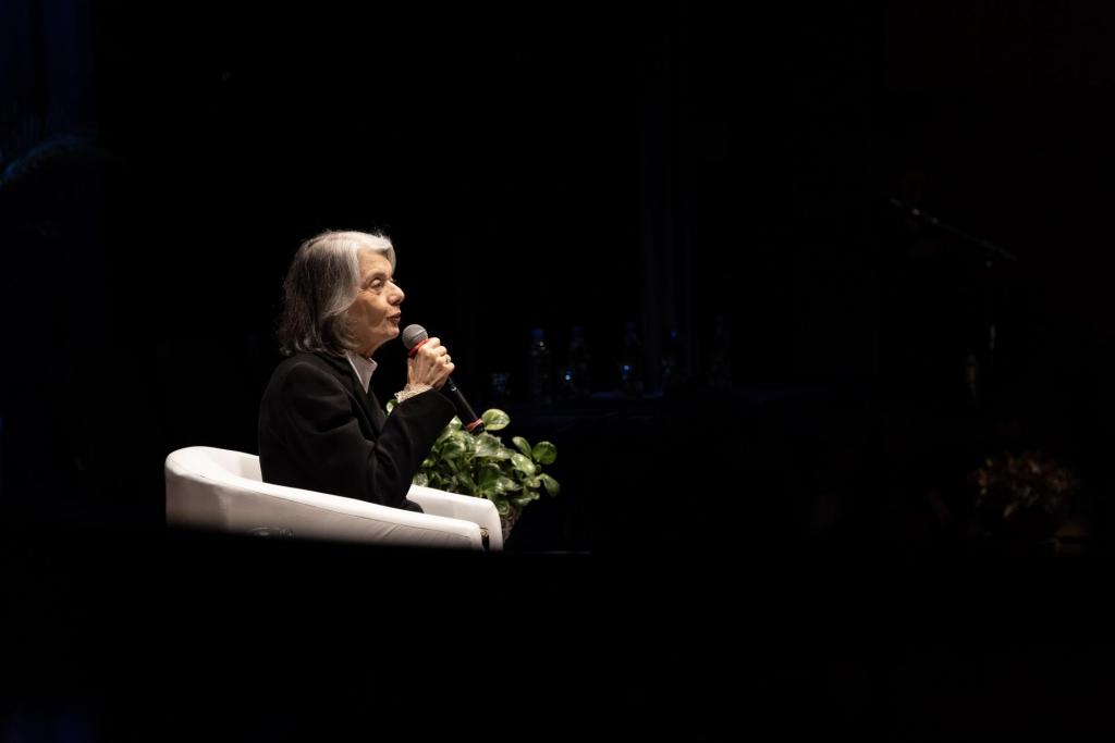 Foto colorida na horizontal da ministra Cármen Lúcia de perfil, à esquerda, falando ao microfone durante palestra. Ela está sentada em uma poltrona branca e mantém a mão perto da boca segurando o microfone enquanto ministra a palestra. Está vestindo roupa preta e com um holofote de luz sobre ela. O Ambiente é escuro.