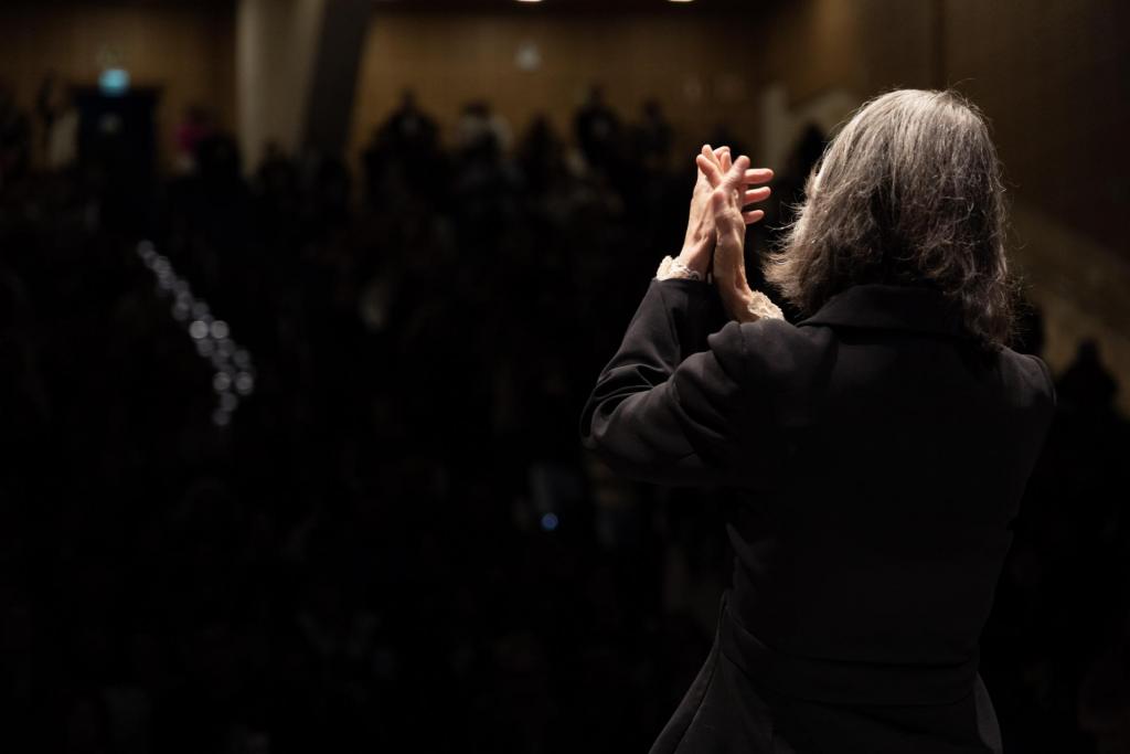 Foto colorida na horizontal, registra a ministra Cármen Lúcia de cabelos grisalhos, vista de costas, diante da plateia durante a cerimônia. Ela veste uma roupa preta e mantém as mãos levantadas, com os dedos entrelaçados, em um gesto de agradecimento ao público. Ao fundo, a plateia aparece sentada em um ambiente escuro, com a iluminação concentrada sobre a mulher e parte das paredes do espaço.