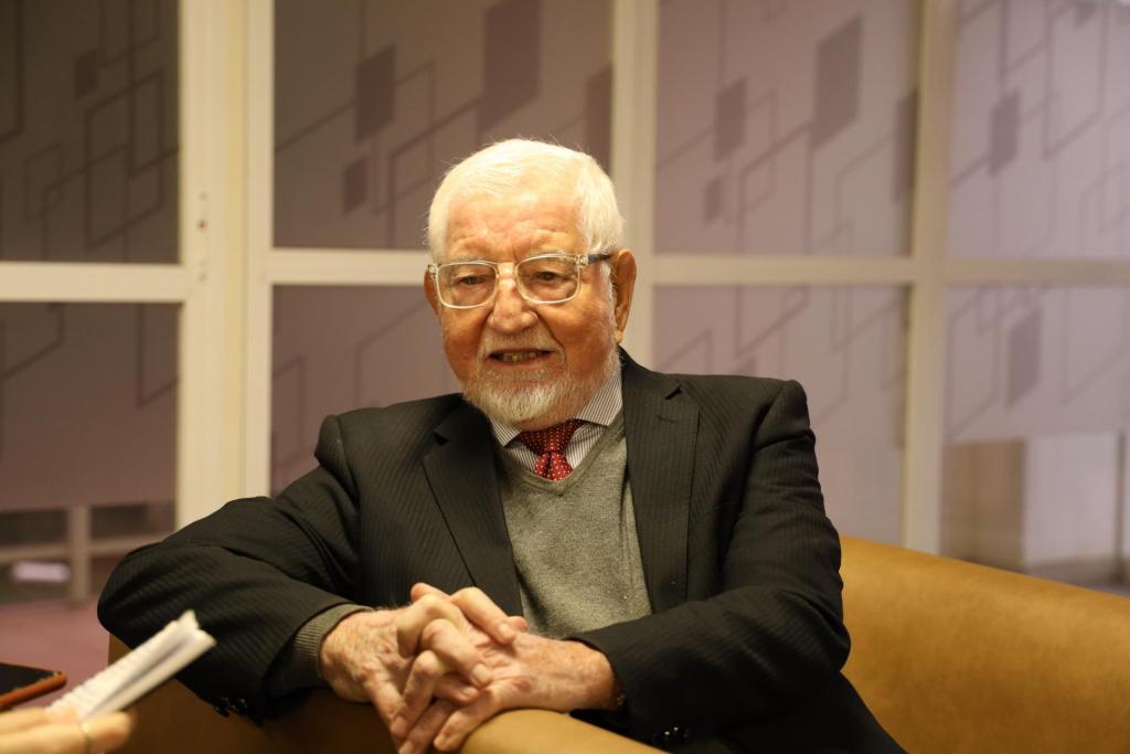 Foto colorida, na horizontal, mostra José Carlos Libâneo durante uma entrevista. Ele está sentado em um sofá, usando terno escuro, camisa, gravata vermelha e suéter cinza. Está com as mãos entrelaçadas sobre o colo e olha em direção à entrevistadora, parcialmente visível no canto inferior esquerdo, segurando um bloco de anotações. Ao fundo, há uma parede com painéis em tons claros e formas geométricas.
