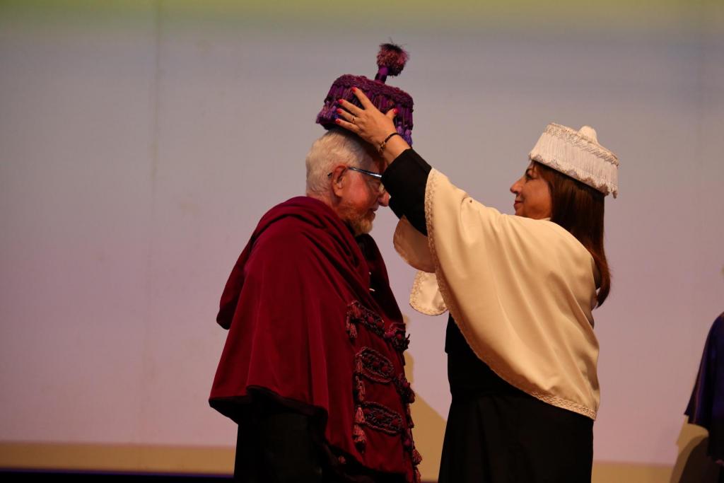 Foto colorida, na horizontal, mostra José Carlos Libâneo e a reitora da UFSM, Martha Adaime, durante a cerimônia de homenagem. Libâneo está à esquerda, de perfil, usando beca vermelha e capelo roxo. Martha está à direita, de frente para ele, colocando o capelo acadêmico sobre sua cabeça com as duas mãos. Ela veste uma beca preta com uma faixa clara sobre os ombros e capelo branco. Ao fundo, há uma tela de projeção em tons claros. O ambiente é iluminado por luzes no palco do Centro de Convenções.