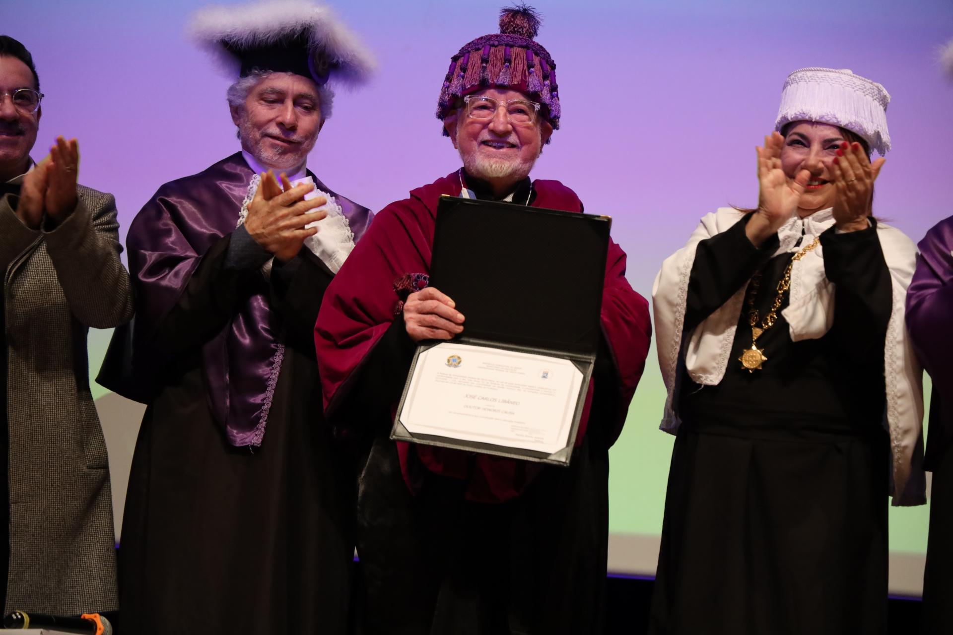 Foto colorida, na horizontal, registra a cerimônia de entrega do doutorado Honoris Causa a José Carlos Libâneo. No centro, Libâneo segura o diploma aberto com um certificado e veste uma beca vermelha, além de um capelo roxo. À esquerda, está o diretor do Centro de Educação da UFSM, Ascísio dos Reis Pereira, parcialmente enquadrado, usando a beca de formatura. À direita, a reitora da UFSM, Martha Adaime, aparece aplaudindo, também com vestimenta acadêmica e capelo branco. Ao fundo, há uma tela iluminada em tons de roxo, azul e verde. O ambiente é iluminado por luzes no palco do Centro de Convenções.