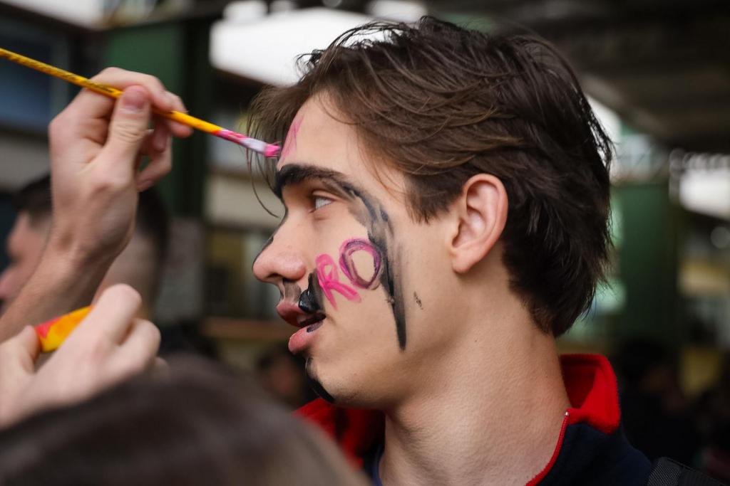 Fotografia colorida na horizontal de um estudante durante uma atividade de
recepção aos ingressantes da Universidade Federal de Santa Maria (UFSM). Em primeiro
plano, no centro da imagem, o estudante aparece de perfil, voltado para a esquerda. Ele
tem cabelos castanhos curtos e veste uma jaqueta escura com gola vermelha. No rosto, há
pinturas em tinta preta e rosa, com um desenho que se estende da testa até a bochecha
esquerda e a palavra "Agro" escrita em letras rosas na lateral do rosto. À esquerda da
imagem, apenas as mãos de outra pessoa aparecem enquanto pintam a testa do estudante
com um pincel de cabo amarelo e tinta rosa. Na parte inferior, outra mão segura um frasco
de tinta nas cores laranja e amarelo. Ao fundo, o ambiente desfocado mostra parte de um
espaço coberto da Universidade, com pilares verdes, luminárias no teto e outras pessoas
reunidas durante a atividade.