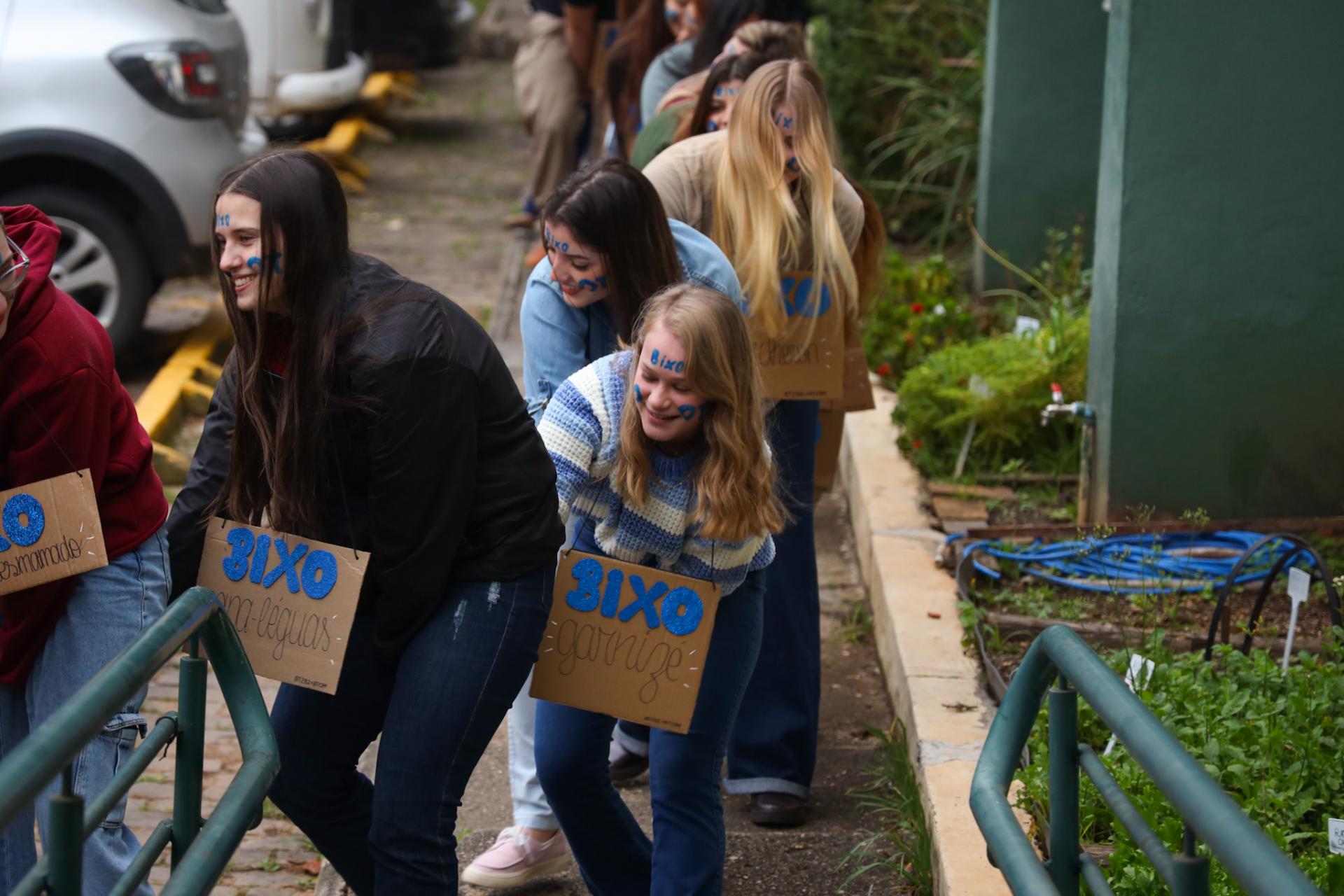 Fotografia colorida na horizontal de uma atividade de integração entre calouros da UFSM. Em primeiro plano, um grupo de estudantes participa de uma dinâmica ao ar livre e organizados em fila. Todos estão levemente inclinados para a frente, com as mãos apoiadas nos ombros da pessoa à frente, enquanto percorrem uma rampa com corrimãos verdes nas laterais. Os estudantes sorriem durante a atividade. No centro da imagem, destacam-se quatro jovens. À esquerda, uma estudante de cabelos longos e escuros veste casaco preto e calça jeans escura. Ela sorri e olha para a frente. Há uma placa de papelão pendurada no pescoço escrita à mão com a palavra "Bixo" em letras azuis e um apelido logo a baixo. Ao lado dela, outra estudante de cabelos escuros usa jaqueta jeans azul-clara, está levemente curvada e olha para a colega à frente. À frente das duas, uma estudante de cabelos loiros na altura dos ombros veste um suéter listrado em azul, branco e cinza e sorri enquanto olha para baixo. Ela também usa uma placa de papelão com a inscrição "Bixo". Mais ao fundo, outra estudante de cabelos longos e loiros cobre parcialmente o rosto com os fios enquanto participa da dinâmica e carrega uma placa semelhante. Os estudantes têm pinturas em tinta azul no rosto, com desenhos e a palavra "bixo" escrita na testa e nas bochechas. Ao fundo, a fila continua com outros participantes, parcialmente encobertos uns pelos outros. À esquerda da imagem aparece parte de um carro branco estacionado. À direita, há um jardim e a parede verde de um prédio