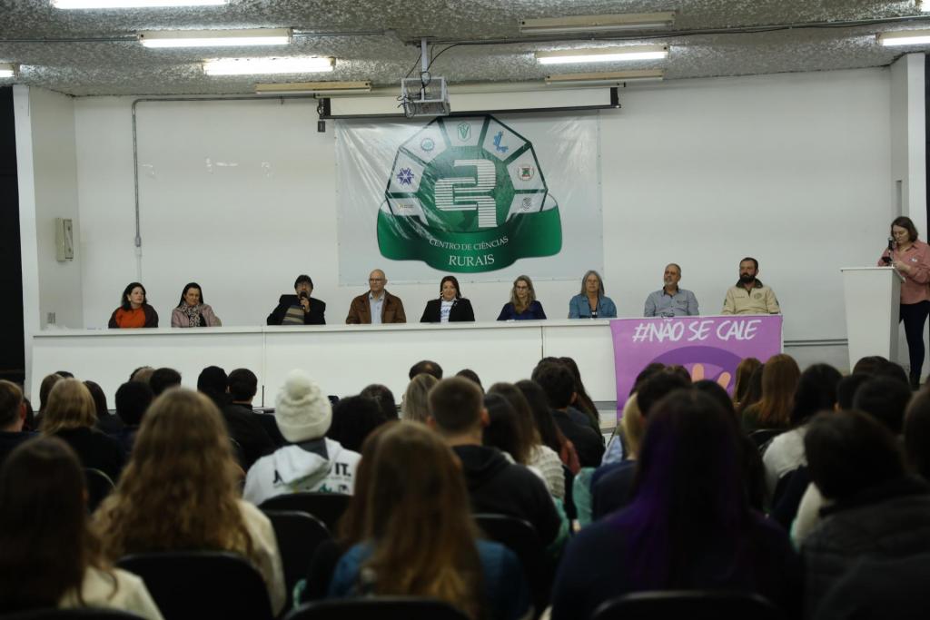 Fotografia colorida na horizontal da recepção aos estudantes ingressantes no
Auditório Flavio Miguel Schneider, do Centro de Ciências Rurais (CCR) da UFSM. Em
primeiro plano, estudantes ocupam fileiras de cadeiras voltadas para o palco. Eles
aparecem de costas e de perfil, vestem roupas de frio em tons escuros e claros e
acompanham a atividade. Ao centro da imagem, uma mesa branca reúne representantes da
Universidade. A reitora Martha Adaime está sentada no centro da mesa, veste blazer preto
sobre camiseta branca e observa a atividade. Ao lado dela, estão os coordenadores dos
cursos do Centro de Ciências Rurais. À esquerda da mesa, um dos coordenadores fala ao
microfone, enquanto os demais permanecem sentados, voltados para o público. À direita do
palco, uma mulher está em pé atrás de um púlpito branco, segurando um microfone. Em
frente ao púlpito, há um painel em tons de roxo e rosa com a frase "#NÃO SE CALE" escrita
em letras brancas. Ao fundo, na parede branca do auditório, há um grande banner com o
símbolo e o nome do Centro de Ciências Rurais, acompanhado dos logotipos das unidades
que compõem o centro. No teto, luminárias fluorescentes e um projetor suspenso iluminam
o ambiente.