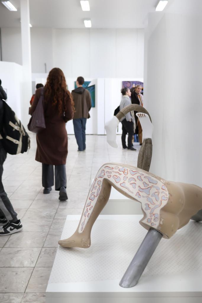 A foto mostra, à direita, uma escultura que faz parte das exposições. Há pessoas circulando pelo ambiente, que tem quadros pendurados nas paredes, as quais são brancas.