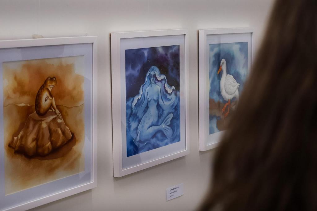 Na foto, aparecem três quadros, pendurados sobre uma parede branca, os quais representam um sapo, uma sereia e um pato (da esquerda para a direita, respectivamente). Em primeira, à direita, aparece uma cabeça desfocada de uma pessoa de costas, a qual tem cabelos compridos, lisos e castanhos.