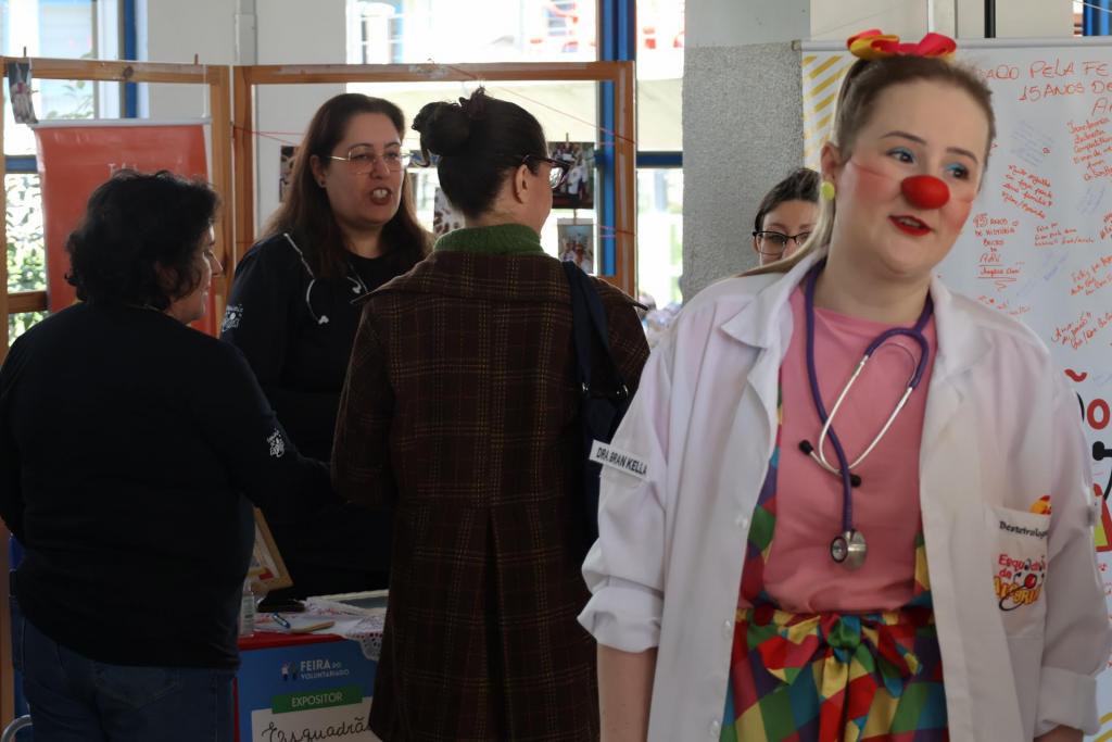 A foto mostra cinco mulheres: duas delas de costas e três de frente, incluindo a mulher branca e loira mais à direita, que se destaca das demais por estar fantasiada de palhaço. A caracterização inclui um nariz redondo vermelho, mas também um estetoscópio pendurado ao pescoço e um jaleco médico na cor branca, sobre o qual (no ombro direito) está escrito o nome da personagem: “Dra. Bran Kella”.