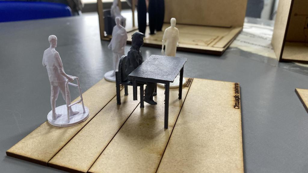 A foto mostra bonequinhos de plástico, nas cores branco e preto, adaptáveis para diferentes ambientes, simulados por meio de peças fabricadas com MDF. Um boneco preto está sentado a uma mesa da mesma cor. Outro boneco, de cor branco, está em pé e com um andador, simulando uma pessoa com dificuldades de locomoção.