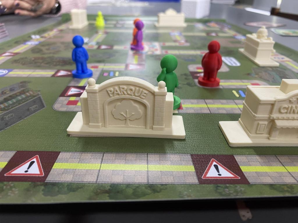 A foto mostra um jogo de tabuleiro com bonequinhos nas cores azul, verde, vermelho, amarelo, laranja e roxo. Sobre o tabuleiro, também estão pequenos displays que simulam locais como parque e cinema, entre outros que podem ser encontrados em uma cidade.