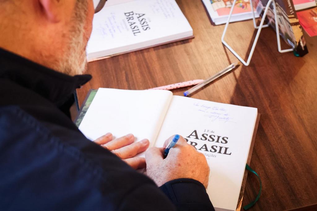 Foto colorida horizontal de homem assinando livro sobre uma mesa. A página do livro traz o nome J.F.Assis Brasil