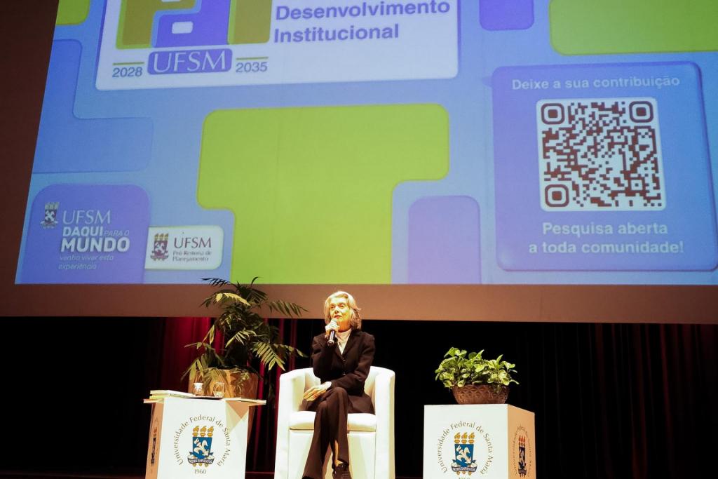 Foto colorida na horizontal que mostra a ministra, ao centro, falando ao microfone na poltrona branca. Ela está com roupa preta e voltada ao público. No seu lado esquerdo e direito, há dois balcões brancos com a logo da UFSM e com plantas em cima. Ao fundo, está projetada uma imagem verde e azul com a logo do novo Plano de Desenvolvimento Institucional e um QR-code à direita para uma pesquisa de opinião pública aberta a toda a comunidade. O ambiente é claro, com iluminação branca no palco e voltada para a ministra.