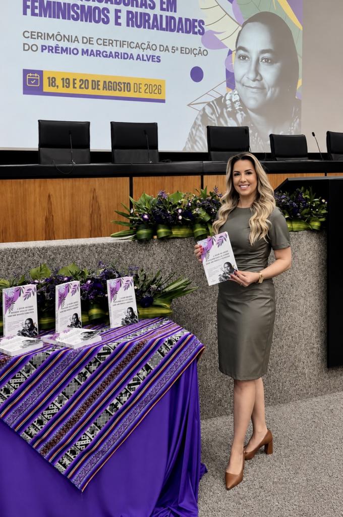 A foto mostra uma mulher branca, de cabelos louros e compridos, que usa sapatos marrons e um vestido de cor verde acinzentado. Ela sorri e segura um livro. Ao fundo, é exibido o cartaz do evento, atrás de uma bancada com microfones e computadores. Ela está em pé, ao lado de uma mesa (coberta com uma toalha roxa), onde são exibidos livros.