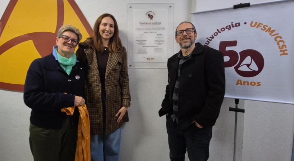 Foto colorida horizontal do descerramento da placa de jubeleu de ouro do curso de Arquivologia. À frente da placa, duas professoras e um professor em pé