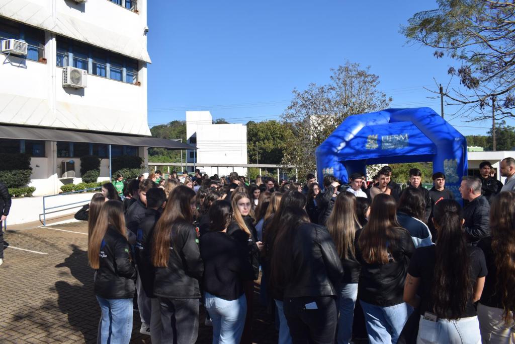 Foto colorida horizontal do campus Frederico Westphalen com um grupo de dezenas de estudantes adolescentes em pé que acompanham a explanação de um homem. Do lado esquerdo deles, um dos blocos do campus. À frente deles, uma item inflável usado em feiras