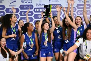 Fotografia colorida horizontal de atletas de basquete feminino comemorando, com os braços erguidos. Ao centro, Bianca Revers ergue um troféu preto e amarelo acima da cabeça, sorrindo, com medalha no pescoço e uniforme azul-marinho com detalhes lilás. Ao redor dela, outras integrantes da equipe comemoram e sorriem, algumas seguram celulares com chamadas de vídeo ativas. Ao fundo, painel publicitário com logotipos de patrocinadores como Estado de Goiás, Goiânia e Citrino.