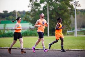 Fotografia colorida horizontal de três pessoas correndo lado a lado em uma pista pavimentada. À esquerda, Gracieli Fernandes corre com cabelos presos em rabo de cavalo, vestindo regata e shorts pretos. Ao centro, o treinador João Vitor Barbieri, o Big John, corre sorrindo, com barba, vestindo regata laranja com a inscrição "Assessoria Big Fort Run" e shorts pretos. À direita, um homem corre vestindo regata e shorts laranja. Ao fundo, gramado, postes e um prédio verde.