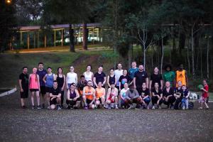 Fotografia colorida horizontal de um grupo de participantes do projeto Corre Junto posando em pé e agachados, em um espaço externo com piso de terra, ao entardecer. Vestem roupas esportivas em tons variados, principalmente preto, com destaque para algumas peças laranja e rosa. À direita do grupo, uma criança de camiseta vermelha segura uma bola e outra criança está agachada ao lado de uma bicicleta infantil azul. Ao fundo, um gramado com árvores altas e, mais ao fundo, um quiosque coberto iluminado.