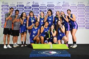 Fotografia colorida horizontal de uma equipe de basquete feminino da UFSM posando para foto após premiação, em pé e agachadas sobre um pódio numerado "3". Todas usam uniforme azul-marinho com detalhes lilás e mordem medalhas penduradas no pescoço. À esquerda, dois homens em pé vestindo camisetas com o logotipo "UFSM Basquete", um deles com o braço apoiado no ombro do outro. Ao fundo, painel publicitário com logotipos de patrocinadores como Estado de Goiás, Goiânia, Spalding e Movida.