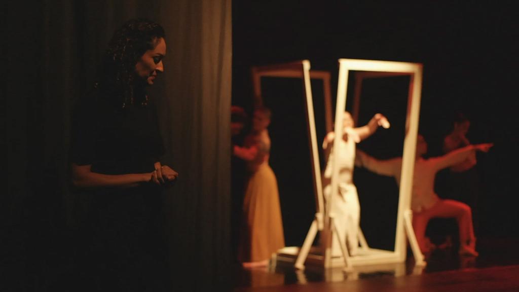 Foto colorida horizontal de grupo de bailarinos no Caixa Preta. No centro da imagem, um bailarino está cercado por pelo menos quadro molduras, que fecham a figura de um quadrado. No entorno, uma bailarina eum bailarino dançam. No canto esquerdo, à frente da cortina, uma mulher observa a cena.