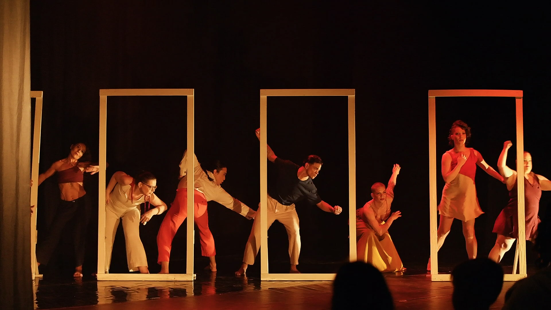 Foto colorida horizontal de grupo de bailarinos no palco do Caixa Preta. As dançarinas e os dançarinos, em pé, fazem coreografias com quatro molduras com tamanho semelhante ao de portas. Alguns estão com os corpos entrando e ou saindo molduras
