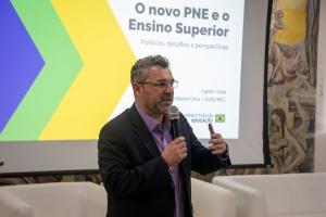 Fotografia colorida horizontal do professor Gregório Grisa falando ao microfone durante uma palestra. Ele está de pé, no centro da imagem, com cabelos e barba grisalhos, óculos de grau, veste camisa lilás e blazer preto. Segura um microfone com a mão direita, próximo à boca, e gesticula com a mão esquerda erguida. Ao fundo, um telão exibe o título "O novo PNE e o Ensino Superior — Políticas, desafios e perspectivas", com o nome "Professor Grisa – SASE/MEC" e o logotipo do Ministério da Educação. À direita, um painel com pintura mural de figuras humanas em tons terrosos e amarelos. À frente, poltronas brancas vazias.