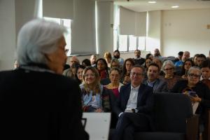 Fotografia colorida horizontal de uma plateia sentada em um auditório, observando a professora Maria Beatriz Luce, que fala de costas para nós, em primeiro plano à esquerda. Ao centro, um homem de cabelos curtos e óculos, vestindo camisa branca e blazer azul-marinho, sorri olhando para ela. Ao lado dele, à esquerda, uma mulher de cabelos loiros e óculos, vestindo jaqueta jeans, também sorri. Atrás e ao redor deles, diversas outras pessoas assistem sentadas, a maioria com expressão atenta. Ao fundo, janelas amplas com cortinas claras e paredes brancas.