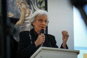 Fotografia colorida vertical da professora Maria Beatriz Luce falando ao microfone em um púlpito. Ela está no centro da imagem, com cabelos brancos curtos, óculos de armação vermelha e batom vermelho. Veste blazer preto sobre blusa estampada com poá branco. Segura um microfone com a mão direita, próximo ao rosto, e ergue a mão esquerda com os dedos unidos, num gesto de explicação. Está apoiada em um púlpito branco. Ao fundo, um painel com pintura mural de uma figura humana em tons terrosos.