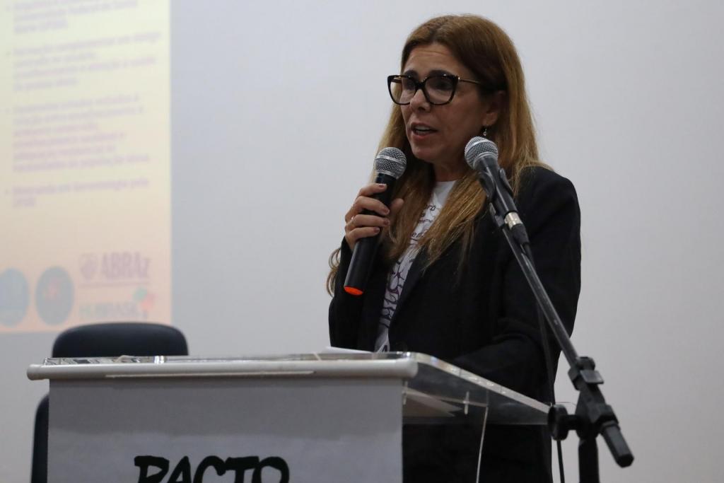 Fotografia colorida na horizontal da professora Kayla Palma, coordenadora do Pacto, em pé atrás de um púlpito. Ela tem cabelos lisos, ruivos, pele clara e usa óculos de armação preta. Veste um blazer preto sobre uma blusa branca com estampa e texto no tecido, além de brincos pequenos e anéis. Segura um microfone de mão preto próximo à boca enquanto fala. Ao fundo, à esquerda, uma tela de projeção com textos e os logotipos da instituição Abraz. Na base do púlpito, a palavra “Pacto” está parcialmente visível.