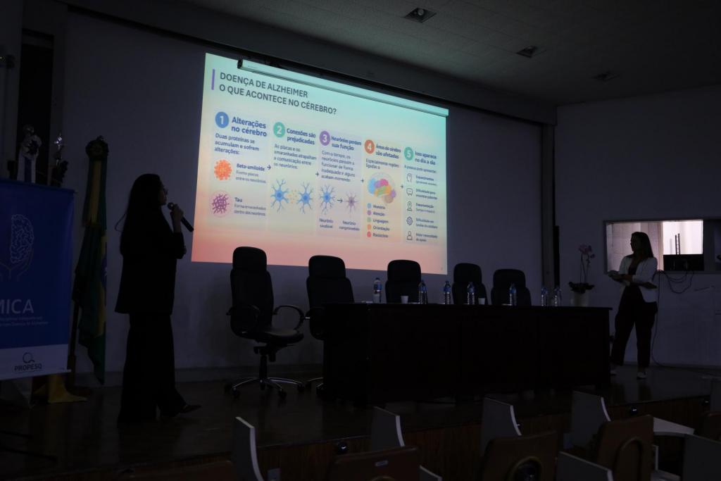 Fotografia do auditório em ambiente escuro durante a apresentação de slides. À esquerda, em silhueta, está Juliana Jornada. Em pé, segura um microfone e fala voltada para a tela de projeção, à direita do palco. Marcielli, também em silhueta, está próxima a uma mesa de apoio com equipamentos. Ao centro, uma mesa vazia com cadeiras pretas e garrafas de água. Na tela de projeção, é exibido um slide com o texto: “Doença de Alzheimer – o que acontece no cérebro?”, organizado em cinco tópicos numerados: “1. Alterações no cérebro: duas proteínas se acumulam e sofrem alterações: Beta-amiloide (forma placas entre os neurônios) e Tau (Forma emaranhados dentro dos neurônios)”; “2. Conexões são prejudicadas: as placas e os emaranhados atrapalham a comunicação entre os neurônios”; “3. Neurônios perdem sua função: com o tempo, os neurônios passam a funcionar de forma inadequada e alguns acabam morrendo” (com ilustração de neurônio saudável e neurônio comprometido); “4. Áreas do cérebro são afetadas: especialmente regiões ligadas à: memória, atenção, linguagem, orientação, raciocínio”; “5. Isso aparece no dia a dia: a pessoa pode apresentar: esquecimentos (geralmente recentes), dificuldade para encontrar palavras, desorientação (tempo e espaço), dificuldade em tarefas do cotidiano, maior necessidade de apoio”.