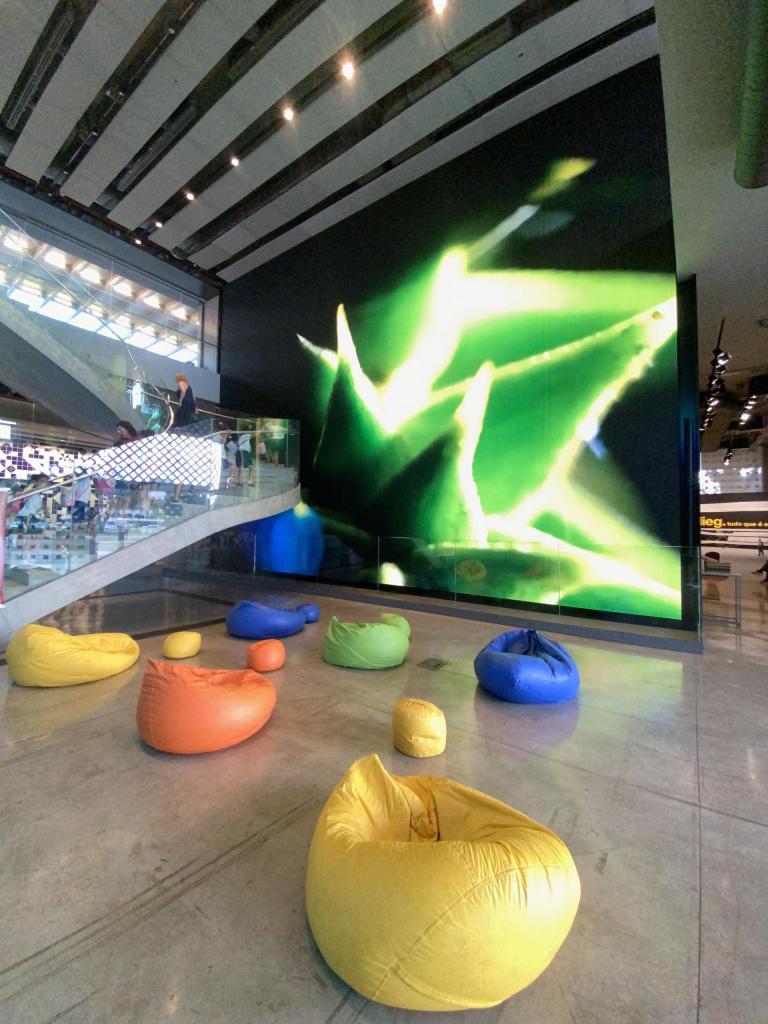 A foto mostra um grande painel de LED localizado atrás de uma escadaria. No chão em frente ao painel, foram colocados vários pufes (de cores variadas), para que o público possa sentar e apreciar as obras audiovisuais exibidas.