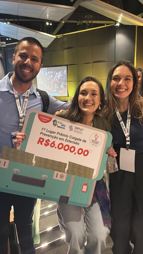 A foto mostra três pessoas em pé: um homem (à esquerda) e duas mulheres. O grupo sorri e segura um cartaz com a inscrição “1º Lugar Prêmio Colgate de Prevenção em Extensão”.