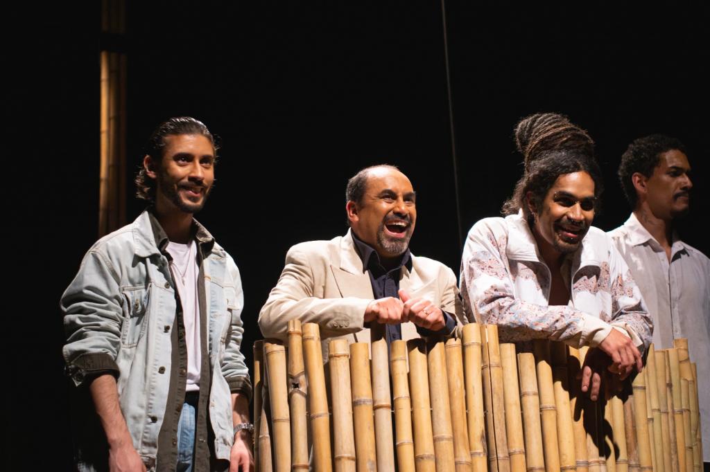 Fotografia colorida, horizontal, em plano médio com iluminação cénica sobre fundo completamente escuro. Quatro homens alinhados lado a lado em um palco de teatro com fundo escuro, três deles estão sorrindo e posicionados atrás de uma estrutura feita com varas de bambu.O primeiro homem tem cabelo escuro e barba curta, veste um casaco jeans claro aberto sobre uma camiseta branca e camisa escura. O segundo homem é calvo, com cabelo e barba grisalhos, veste blazer claro sobre uma camisa escura. Encontra-se atrás da cerca de bambu, debruçado para a frente com as mãos apoiadas e com uma expressão de gargalhada entusiástica. O terceiro homem tem dreadlocks volumosos apanhados no topo da cabeça e barba curta, veste casaco com padrões e encontra-se debruçado sobre a cerca de bambu com os braços cruzados no topo, sorrindo abertamente para a frente. O quarto homem tem cabelo curto e bigode, vestindo uma camisa branca de botões, e está posicionado ligeiramente mais ao fundo.