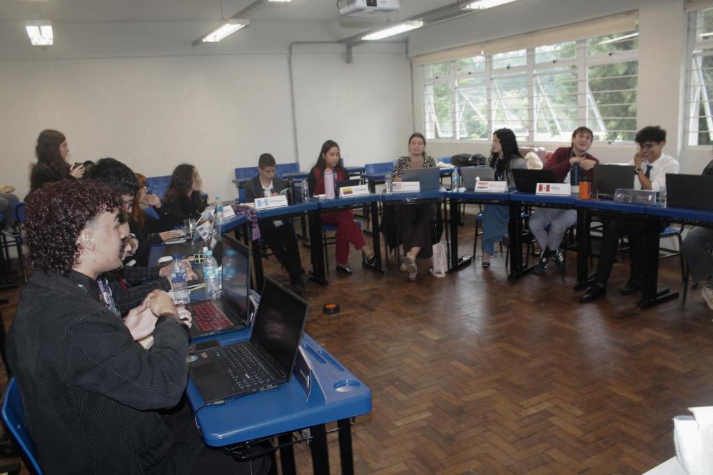 Cerca de 15 alunos estão sentados às mesas da sala de aula, arranjadas em formato de semicírculo. Cada estudante representa um país específico, identificado com uma bandeira sobre a classe.