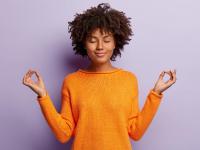 pleasant-looking-calm-woman-meditates-indoor-holds-hands-mudra-gesture-has-charming-smile-closed-eyes-wears-orange-clothes-models-purple-wall-hand-gesture-meditation-concept