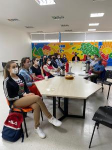 Fotografia colorida na qual 13 pessoas estão ao redor de uma mesa, olhando para o mesmo lado