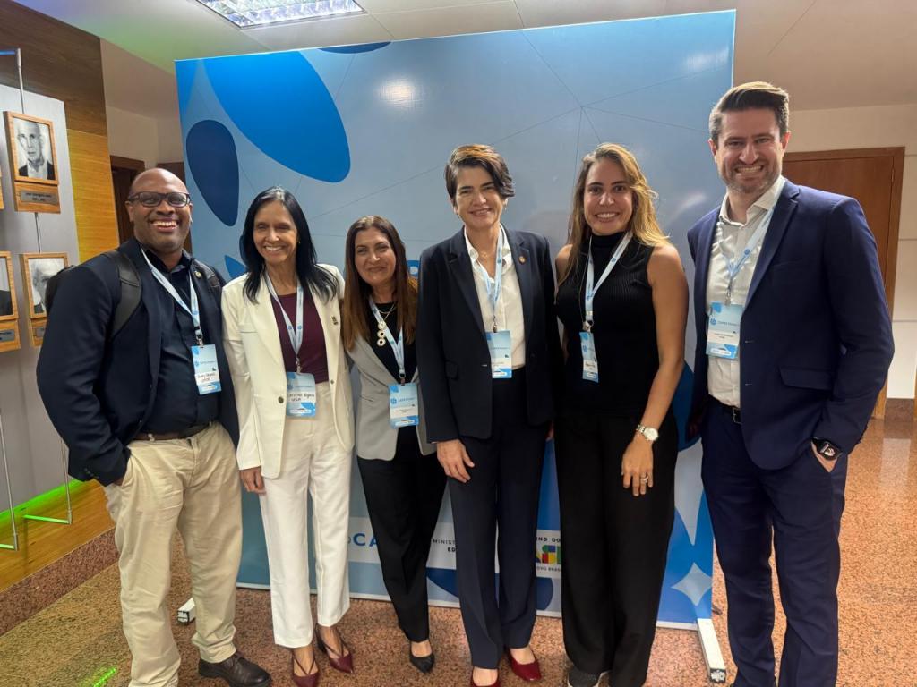 Registro fotográfico com a presença de seis representantes da proposta coordenada pela UFPE. Ambos estão posicionados de pé, um ao lado do outro. Em meio a eles, os professores Tiago Bandeira Marchesan (Vice-Reitor) e Cristina Wayne Nogueira (Pró-Reitora de Pós-Graduação e Pesquisa). O registro foi feito em uma salão aberto em tons marrons amadeirados e com um painel azul ao fundo, com desenhos de algumas pétalas em vários tons de azul.