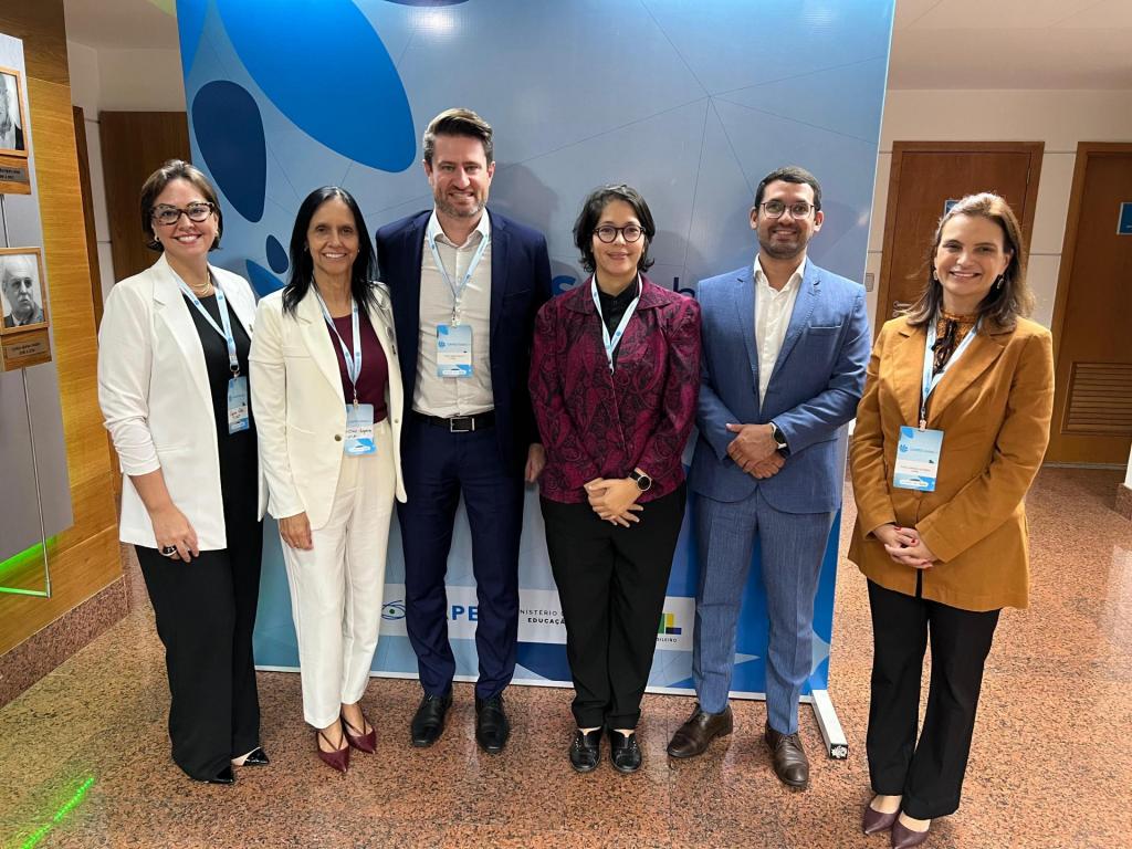 Registro fotográfico com a presença de seis representantes da proposta coordenada pela UFSM. Ambos estão posicionados de pé, um ao lado do outro. Em meio a eles, os professores Tiago Bandeira Marchesan (Vice-Reitor) e Cristina Wayne Nogueira (Pró-Reitora de Pós-Graduação e Pesquisa). O registro foi feito em uma salão aberto em tons marrons amadeirados e com um painel azul ao fundo, com desenhos de algumas pétalas em vários tons de azul.