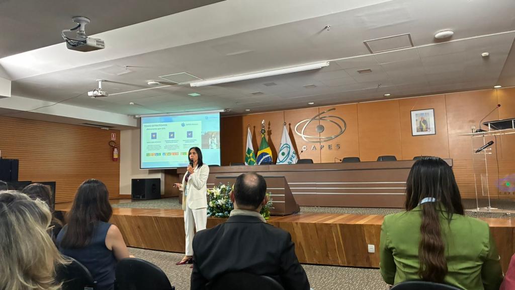 Pró-Reitora de Pós-Graduação e Pesquisa, Professora Cristina Wayne Nogueira fala durante o evento Marco Zero Capes Global no auditório da CAPES em Brasília. No fundo da foto, um painel de madeira com a logo da CAPES no centro, em composição com as bandeiras de Brasília, do Brasil e da CAPES, nesta sequência, localizadas no canto esquerdo, e uma projeção realizada em um retângulo de lona com uma apresentação em slides.