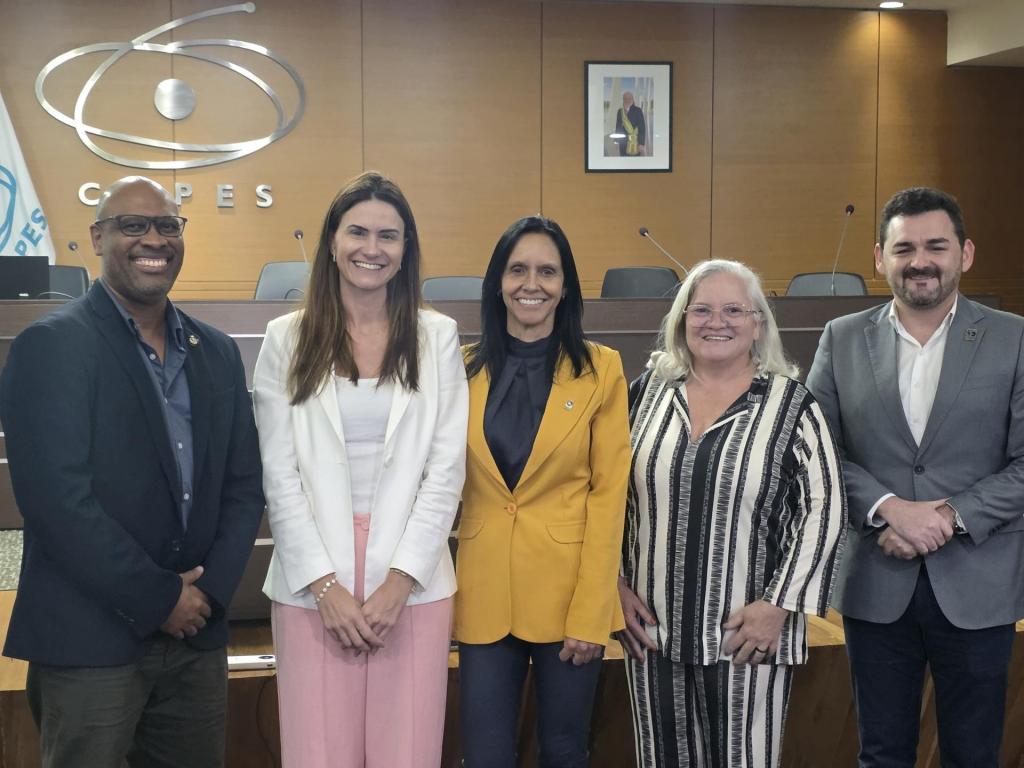 Fotografia com dois homens e três mulheres, lado a lado, sorrindo, presentes Silvana Zucolotto (UFRN), presidenta do colegiado e representante da região Nordeste, Ronney Arismel Mancebo Boloy (CEFET/RJ), pela região Sudeste, Kênia Ferreira Rodrigues (UFNT), pela região Norte e Cristina Wayne (UFSM) pela região sul. Em um auditório, com fundo de madeira, e logo da CAPES.