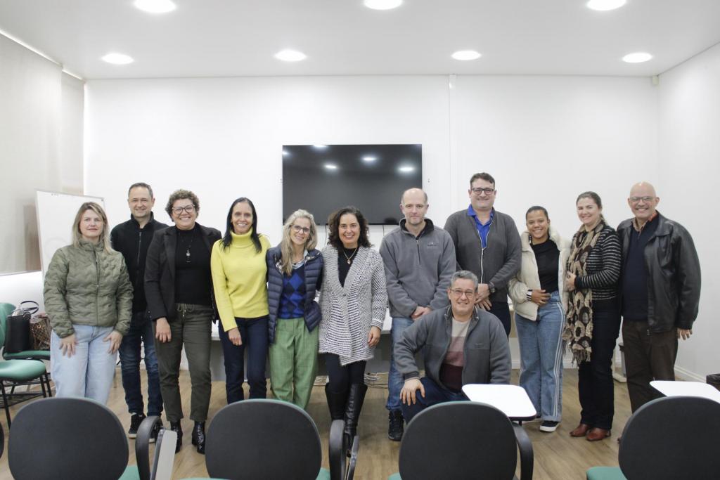 Fotografia de grupo em uma sala de reuniões. Cerca de 12 pessoas posam diante de uma televisão fixada na parede. Uma pessoa está agachada à frente e as demais permanecem em pé. O ambiente possui iluminação embutida e cadeiras de auditório em primeiro plano.