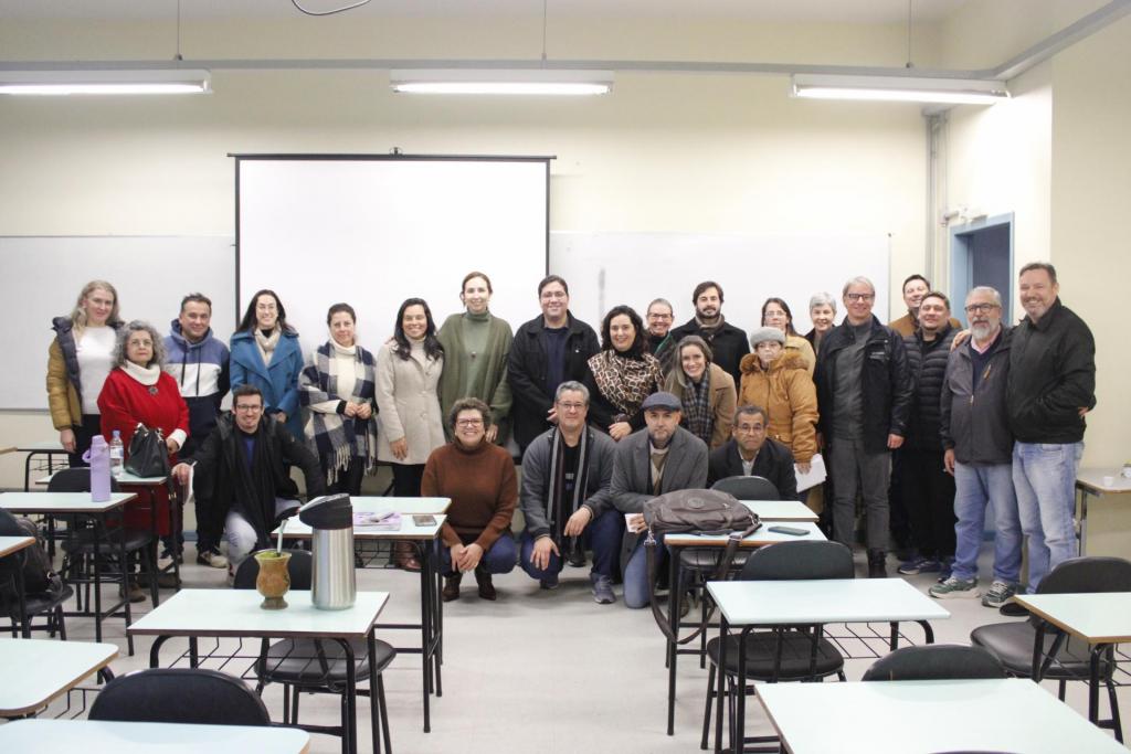 Fotografia de grupo em uma sala de aula. Cerca de 25 pessoas posam para a câmera entre mesas e cadeiras escolares, em frente a um quadro branco e uma tela de projeção. Algumas estão em pé e outras agachadas na primeira fila. O ambiente é iluminado por luzes fluorescentes e os participantes usam roupas de inverno.