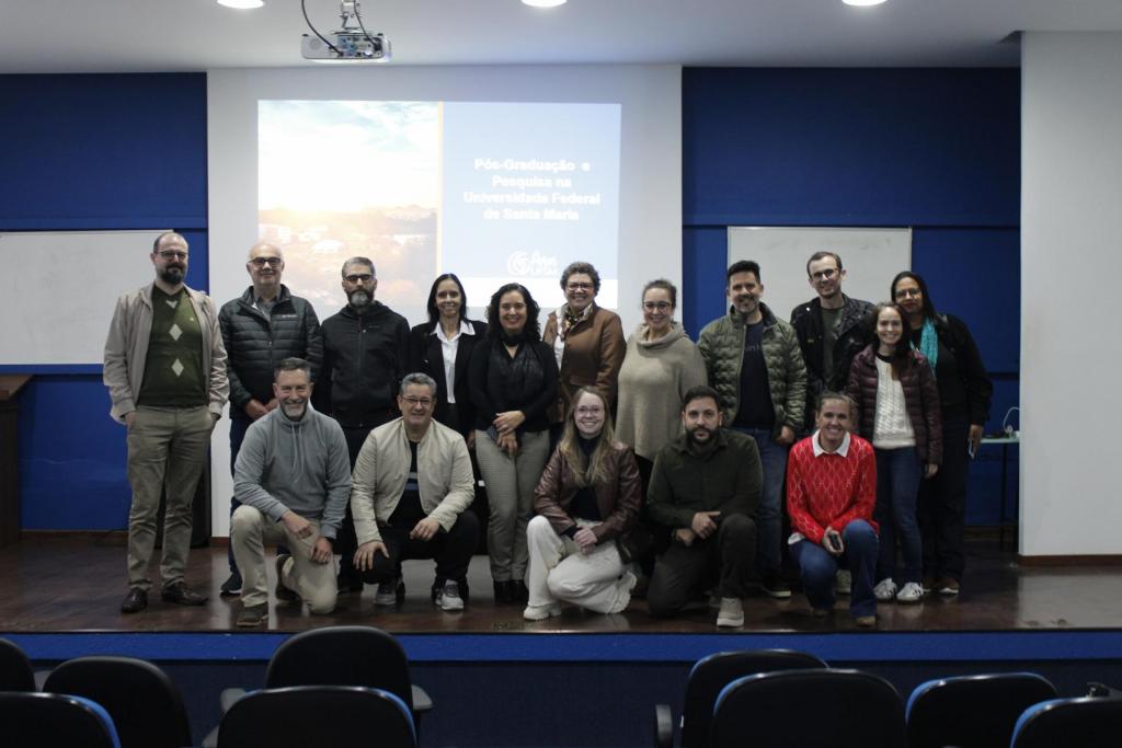 Fotografia de grupo em um auditório. Cerca de 20 pessoas posam sobre o palco em frente a uma tela de projeção que exibe a apresentação "Pós-Graduação e Pesquisa na Universidade Federal de Santa Maria". Parte do grupo está em pé e parte agachada na primeira fila.