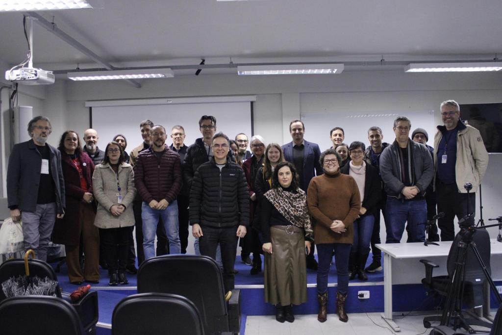 Grupo de cerca de vinte e cinco pessoas reunidas em auditório do CTISM, posando para uma fotografia. A maioria está em pé em diferentes fileiras, enquanto duas pessoas estão posicionadas à frente. O ambiente possui projetor no teto, tela de projeção ao fundo, mesas, cadeiras e equipamentos de gravação, como câmeras e tripés, visíveis no lado direito da imagem. Todos estão voltados para a câmera, usando roupas de frio.