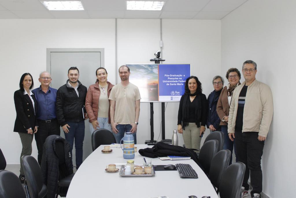Fotografia de grupo em uma sala de reuniões. Dez pessoas posam ao lado de uma tela que exibe uma apresentação sobre pós-graduação e pesquisa na Universidade Federal de Santa Maria. À frente há uma mesa de reuniões com xícaras, garrafa térmica, teclado e outros materiais.