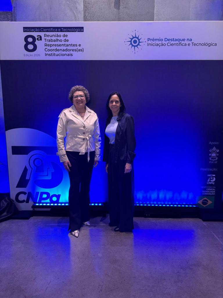 Fotografia de um evento institucional em um espaço interno, com iluminação predominantemente azul. Ao fundo, há um painel com as inscrições “Iniciação Científica e Tecnológica – 8ª Reunião de Trabalho de Representantes e Coordenadores(as) Institucionais – Edição 2026” e “Prêmio Destaque na Iniciação Científica e Tecnológica”, além dos logotipos do CNPq e das instituições apoiadoras. Em frente ao painel, duas mulheres posam lado a lado para uma fotografia. Ambas estão de pé e sorrindo para a câmera, vestidas com roupas sociais. O ambiente tem paredes de tijolos aparentes e piso claro. Posando para a foto encontram-se a Pró-Reitora de Pós-Graduação e Pesquisa, Prof.ª Cristina Wayne Nogueira, e a Coordenadora de Pesquisa, Prof.ª Cristiane de Bona da Silva.