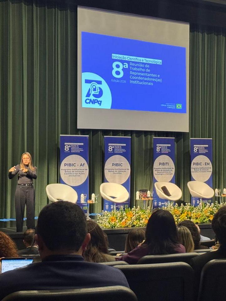 Fotografia de um auditório durante a 8ª Reunião de Trabalho de Representantes e Coordenadores(as) Institucionais de Iniciação Científica e Tecnológica, edição 2026. Ao fundo, um telão exibe a identidade visual do evento. No palco, há quatro painéis verticais com informações sobre programas de iniciação científica, incluindo PIBIC-Af, PIBIC, PIBITI e PIBIC-EM. Uma intérprete de Libras está posicionada à esquerda do palco, enquanto uma pessoa fala ao púlpito à direita. O palco possui cadeiras, pequenas mesas, troféus e arranjos florais. Na parte inferior da imagem, vê-se o público sentado no auditório, observado de costas.