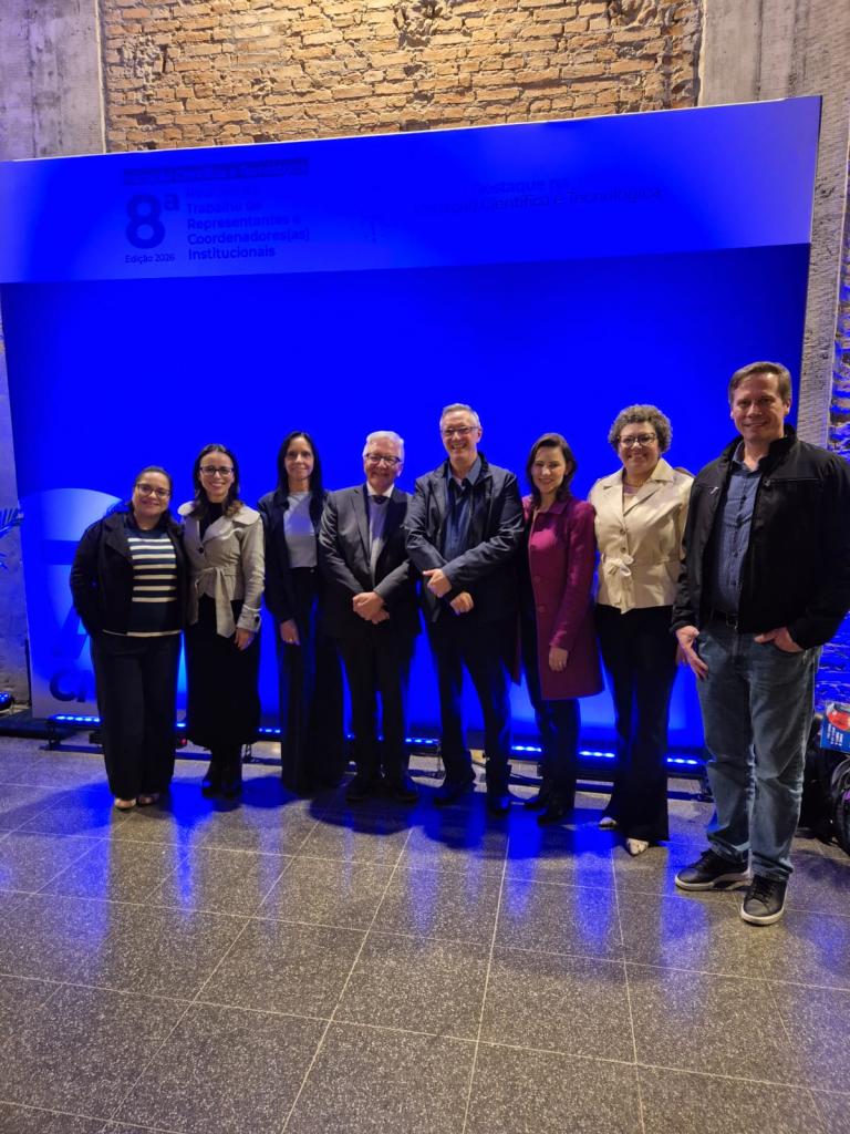 Fotografia de um grupo de oito pessoas reunidas em frente ao painel oficial da 8ª Reunião de Trabalho de Representantes e Coordenadores(as) Institucionais de Iniciação Científica e Tecnológica – Edição 2026. O grupo está de pé, lado a lado, posando e sorrindo para a câmera. As pessoas vestem roupas predominantemente sociais e casuais formais. O fundo é iluminado em azul e apresenta o logotipo do CNPq e a identidade visual do evento. O espaço possui piso escuro e paredes com tijolos aparentes.