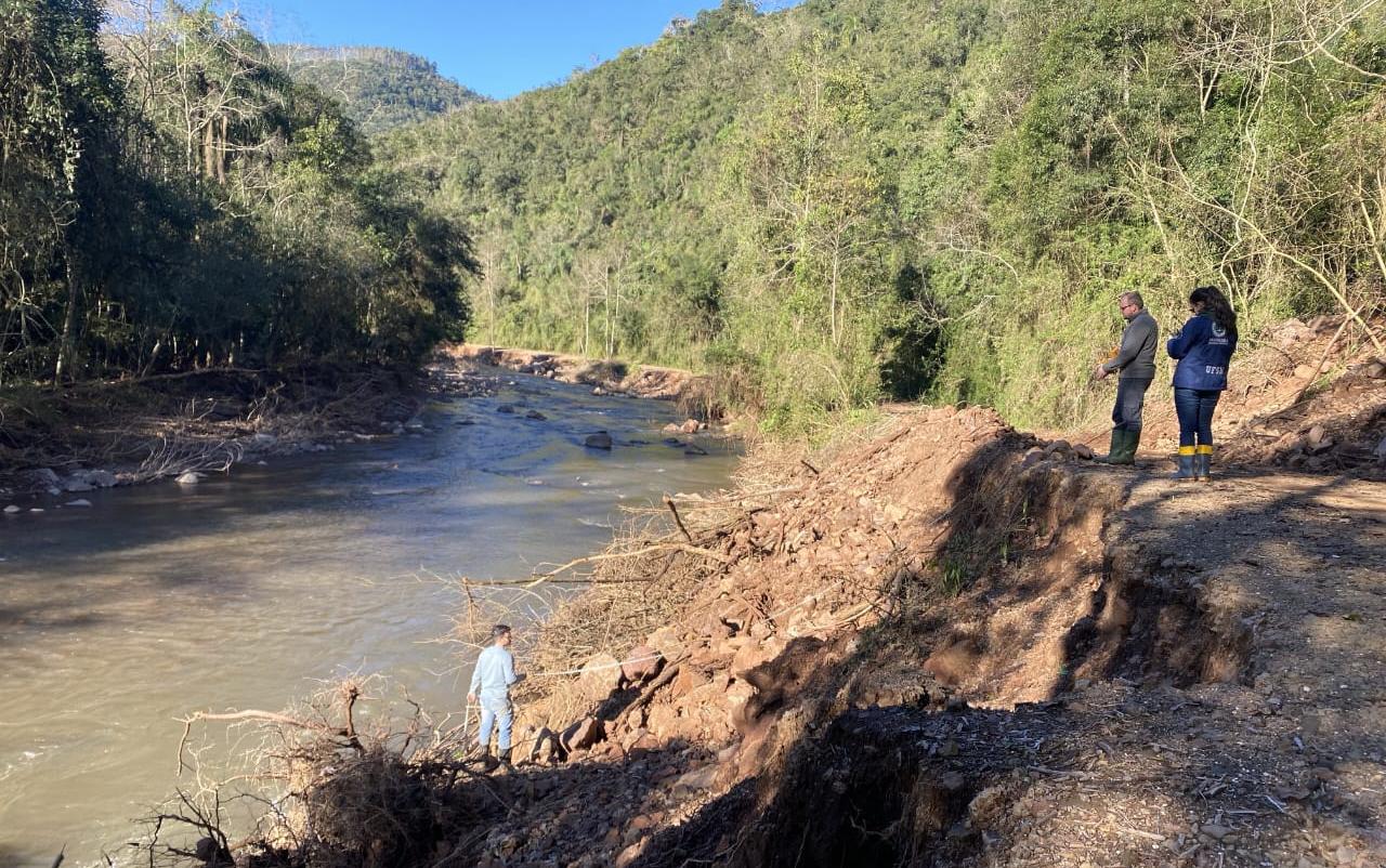 Foto colorida de equipe de engenharia em visita no município de Sinimbu. Na imagem aparecem duas pessoas no canto direito olhando em direção ao rio, que aparece no lado esquerdo da imagem. Uma pessoa está mais próxima ao córrego de água. Ao fundo, árvores e montanhas